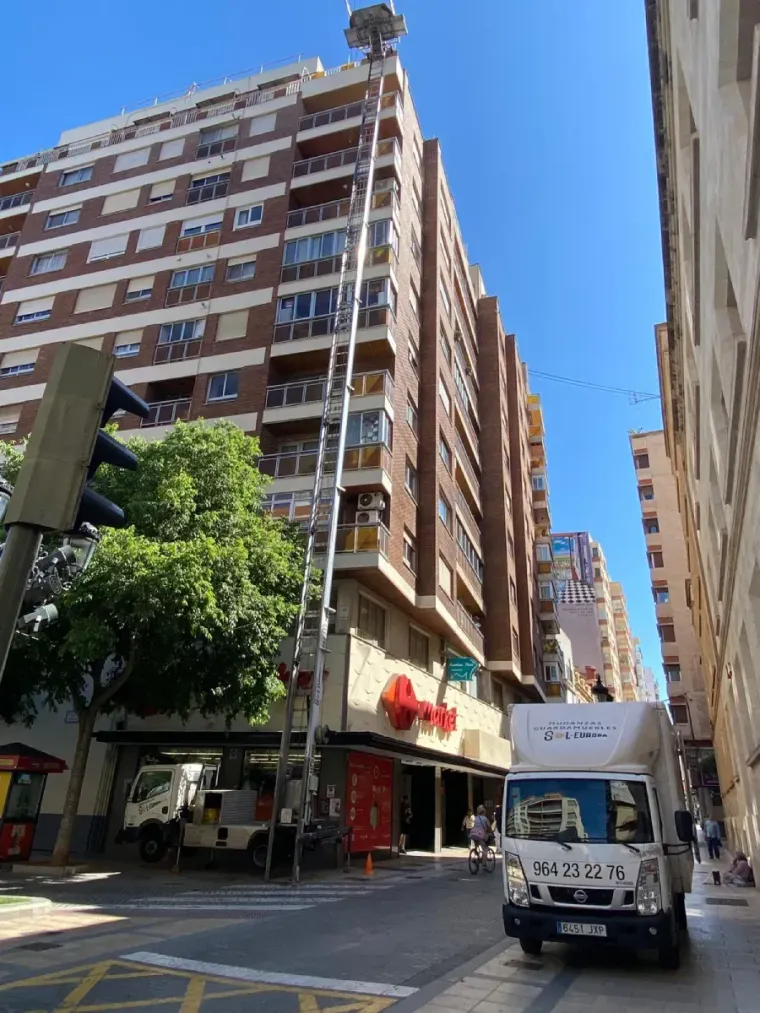 Camión de mudanzas con elevador extendido al lado de un edificio alto, cielo azul.