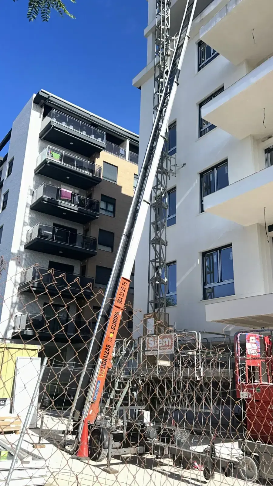 Un ascensor naranja y blanco que sube por un edificio. Obras con vallado.