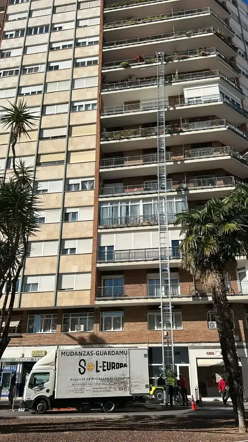 Camión de mudanzas con elevador que se extiende a un edificio de varios pisos para entrega de muebles.
