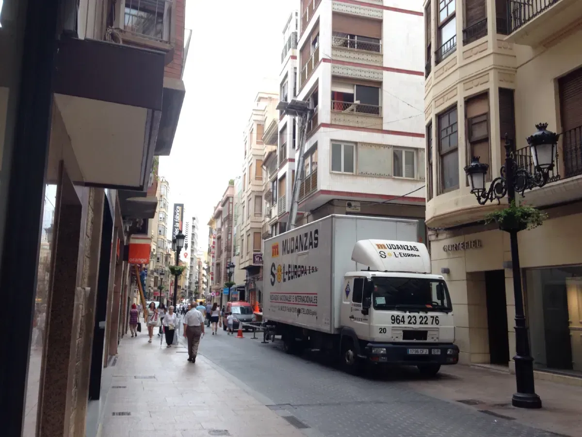 Camión en una calle estrecha en Valencia, España, peatones caminando por las aceras, edificios altos a ambos lados.