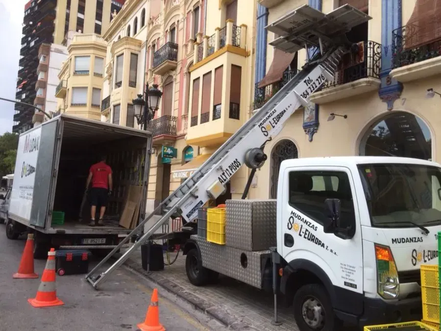 Camión descargando muebles con un elevador extensible en una calle de la ciudad. Trabajadores trasladando objetos.