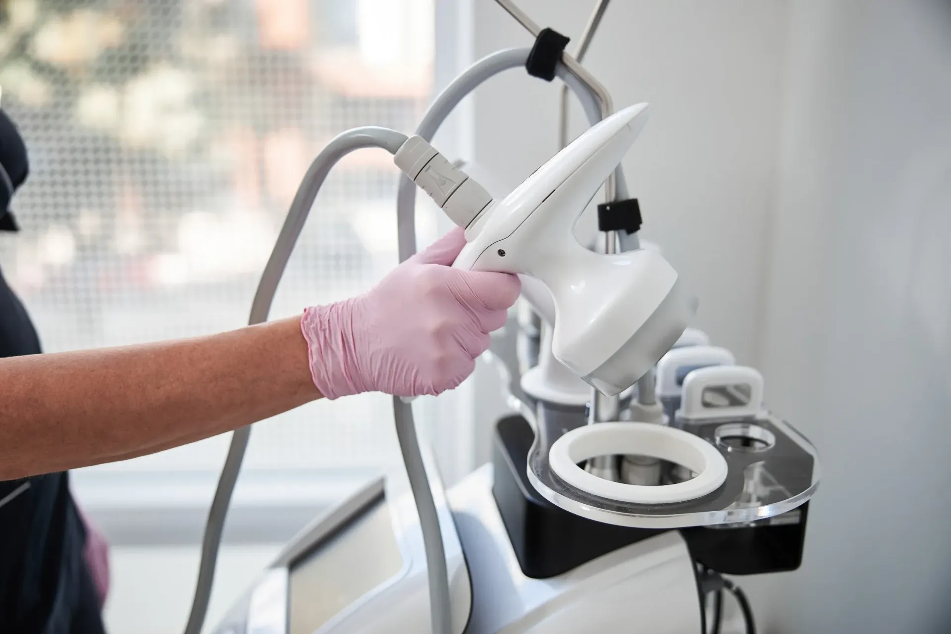 Mano enguantada que sostiene un dispositivo cosmético blanco y gris; se ven tubos y controles.