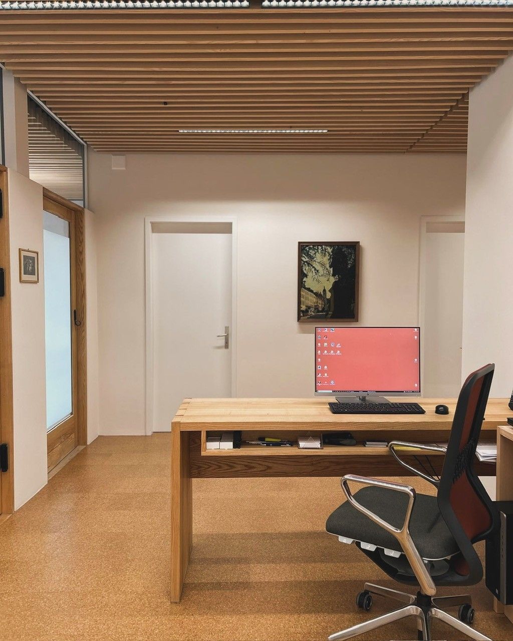 Büroraum: Holzschreibtisch, Computer, schwarzer Stuhl, Holzdecke, weisse Wände, Korkboden.