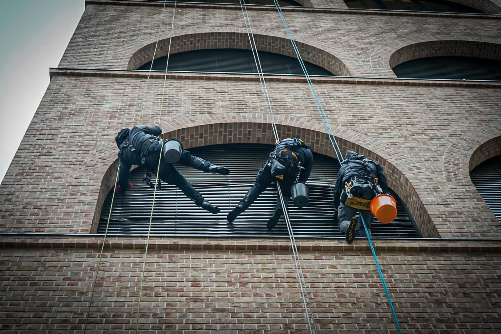 Cleanclimb: Eine Gruppe von Mitarbeiter hängen an einem Backsteingebäude um die Jalousien am Fenster zu reinigen.