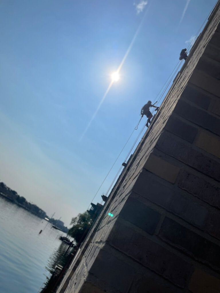 Cleanclimb: Ein paar Mitarbeiter hängen an einem Gebäude und reinigen die Fassade am Rande eines Flusses. 