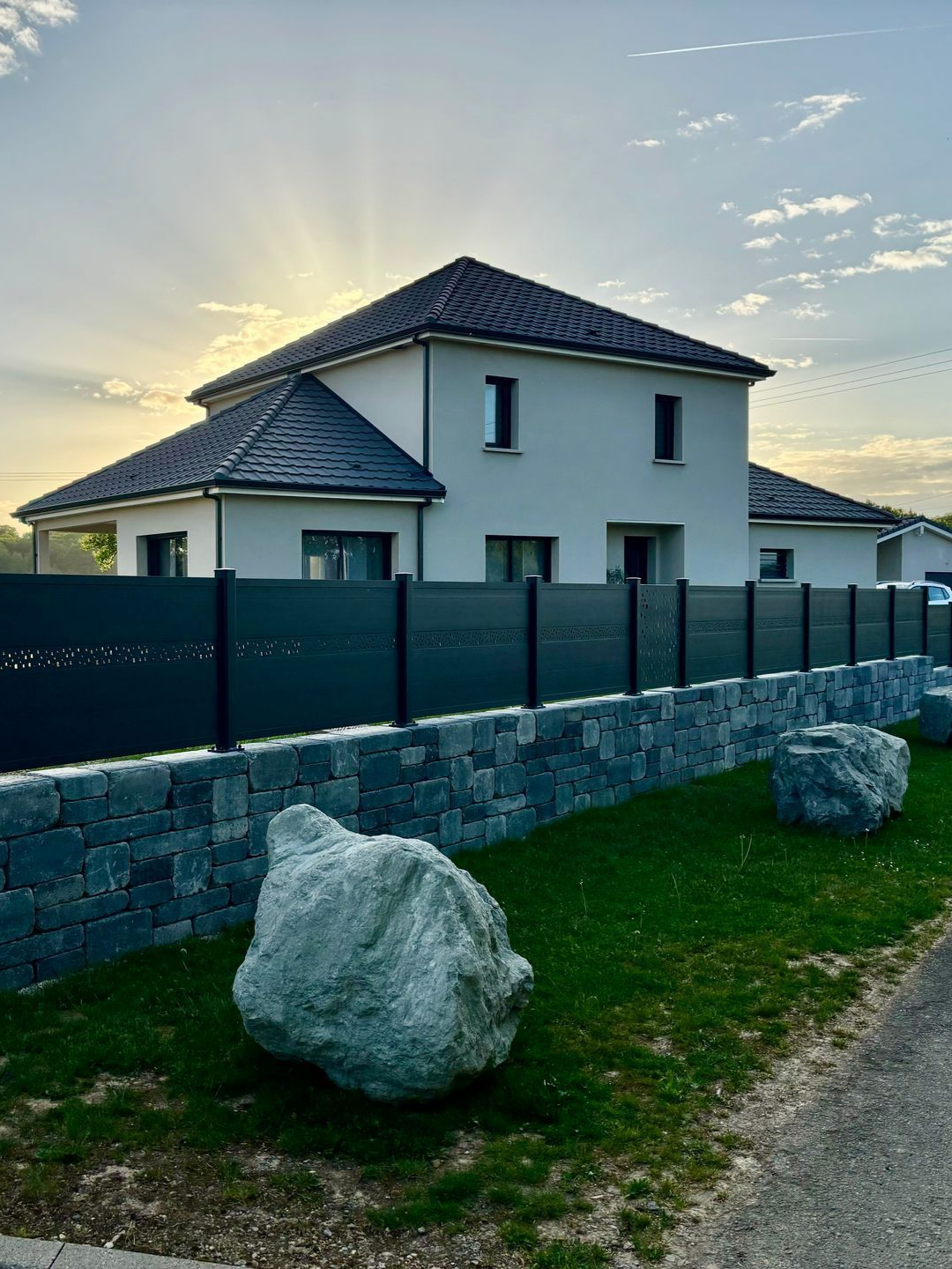 Maison blanche moderne derrière une clôture sombre et un mur de pierre. De gros rochers jonchent la pelouse.