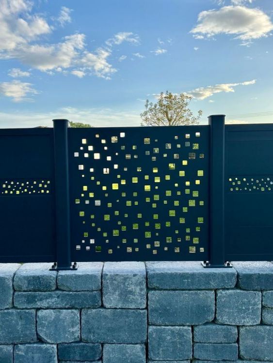 Clôture en métal noir avec des découpes carrées sur un mur de pierre sous un ciel nuageux.