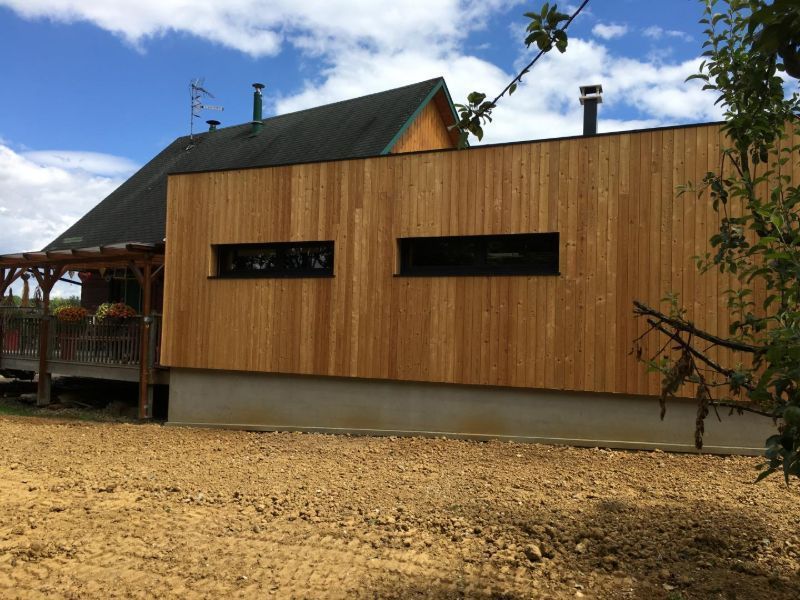 Maison à façade en bois avec petites fenêtres rectangulaires, construite sur des fondations en béton, ciel bleu nuageux.