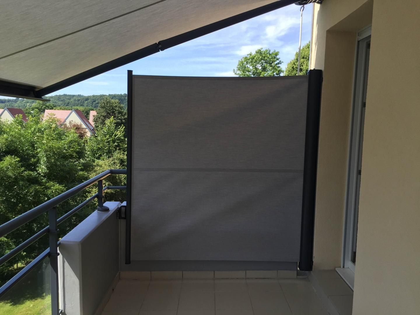 Store vertical gris de type paravent rétractable sur balcon, donnant sur les arbres et les maisons.