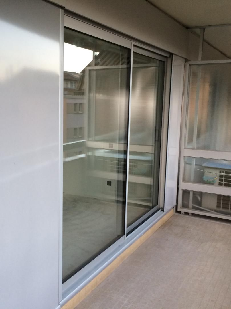 Porte-fenêtre coulissante en verre, cadre en aluminium, partiellement ouverte, donnant sur un balcon. Murs gris, sol clair.