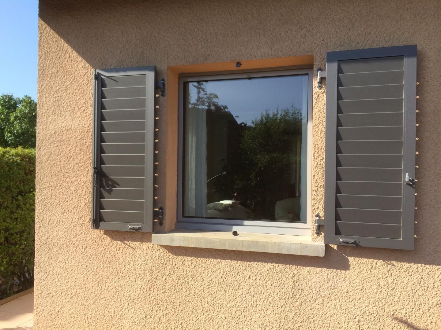 Fenêtre à volets gris sur un mur en stuc couleur pêche ; les volets sont ouverts.