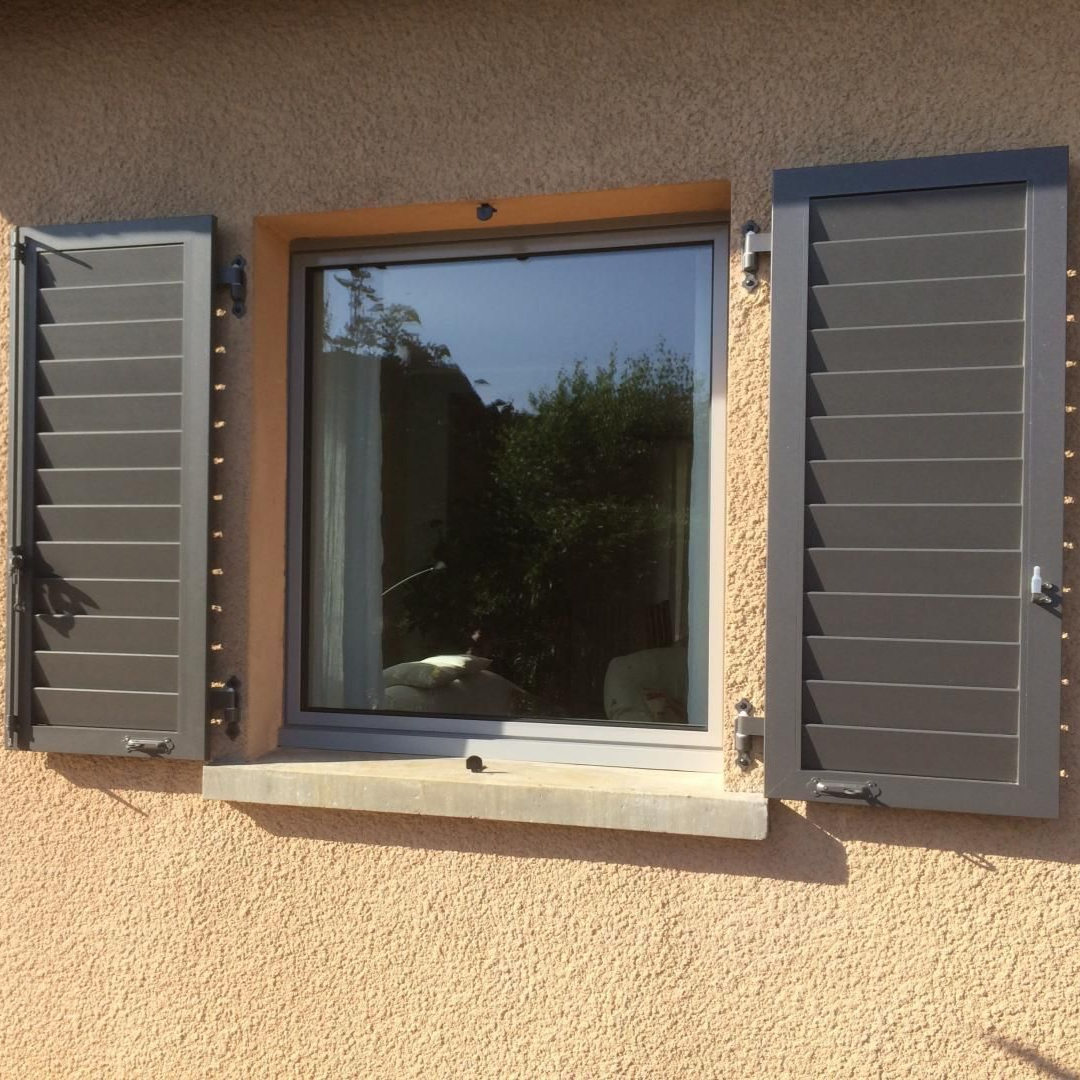 Fenêtre à persiennes grises ouvertes sur un mur en stuc beige ; un jardin se reflète dans la vitre.