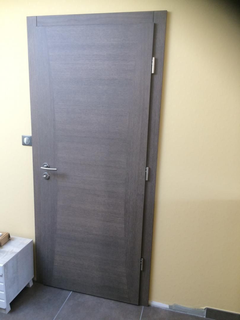 Porte en bois gris contre un mur jaune, avec une poignée et des charnières argentées.