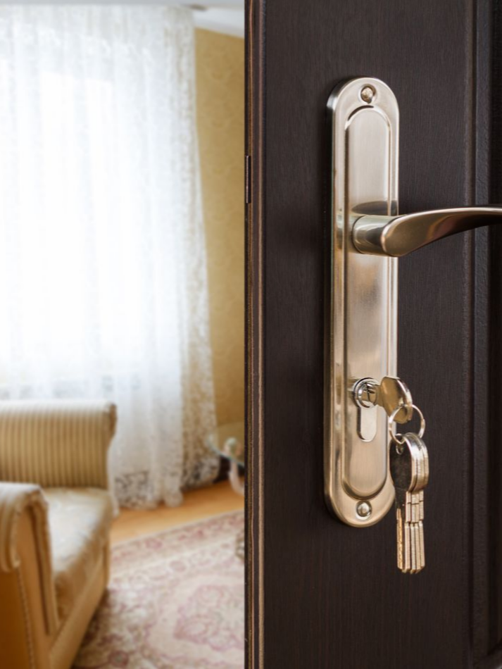 Une porte noire ouverte, munie d'une poignée et de clés argentées, révèle un salon avec un canapé et une fenêtre.