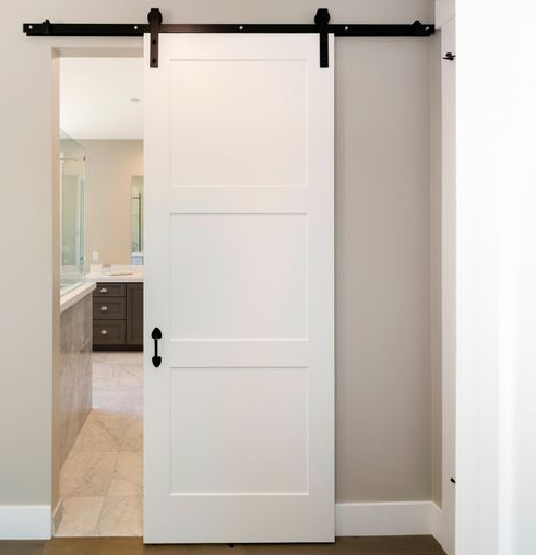 Porte à galandage blanche, quincaillerie noire, ouverte sur une salle de bains avec armoires grises et baignoire.