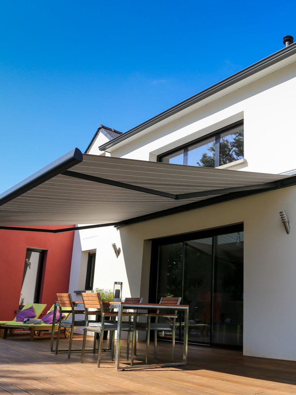 Terrasse extérieure avec store rétractable, table, chaises et une maison blanche sous un ciel bleu.