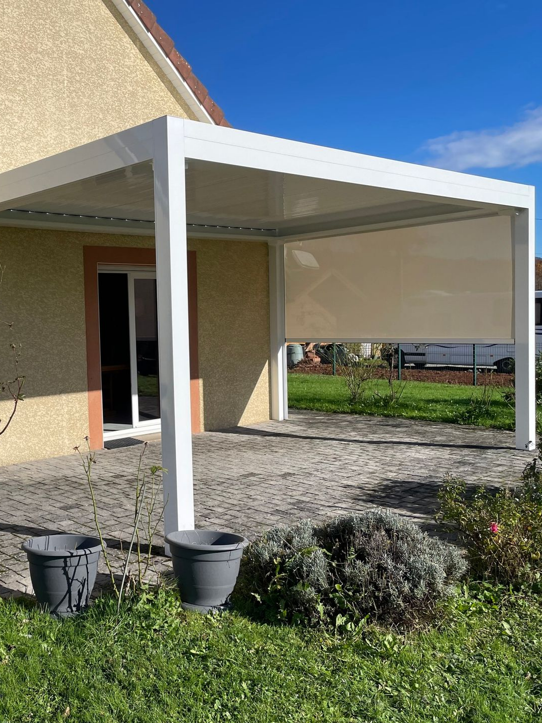 Pergola blanche adossée à une maison beige, avec un store enroulé.