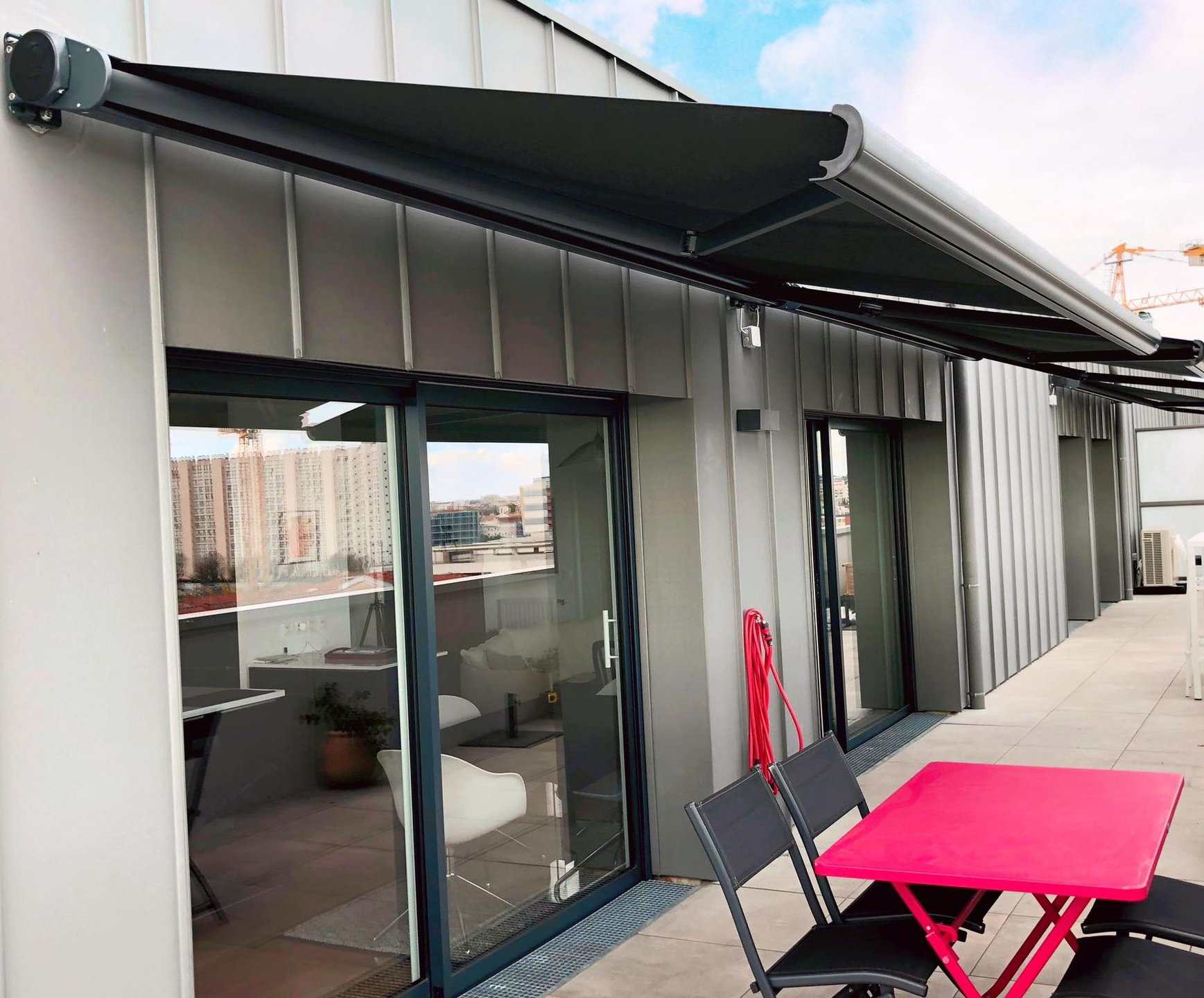 Store banne rétractable gris au-dessus d'un balcon avec portes vitrées, table rose et chaises.