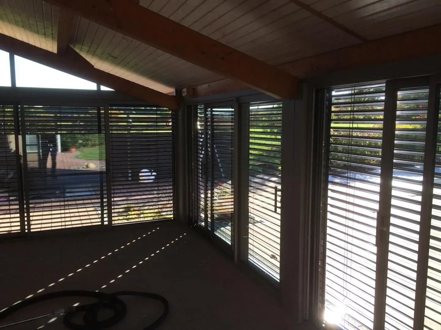 Intérieur d'une véranda avec stores à lamelles fermées, poutres en bois et vue sur l'extérieur.