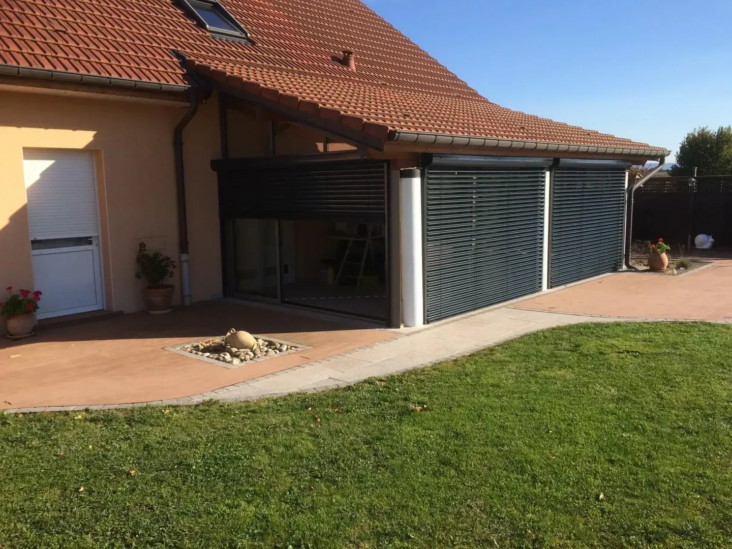 Vue extérieure d'une maison avec un toit de tuiles, des murs beiges et une véranda avec volets sombres fermés.
