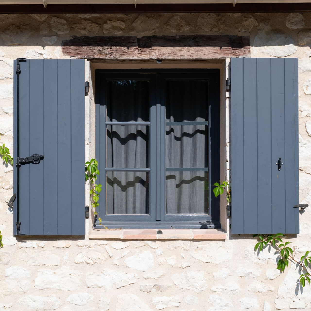 Fenêtre à volets gris sur un mur de pierre, avec des vignes et un cadre en bois sombre.