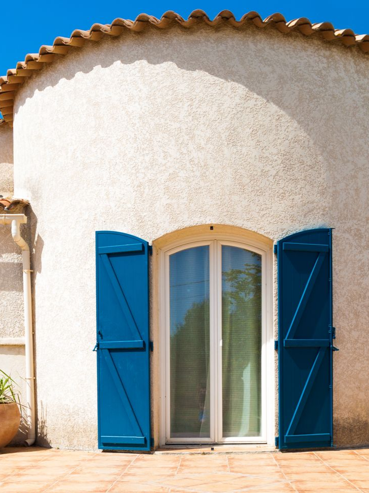 Bâtiment en stuc blanc avec fenêtre cintrée, volets bleus et toit de tuiles en terre cuite sur fond de ciel bleu.