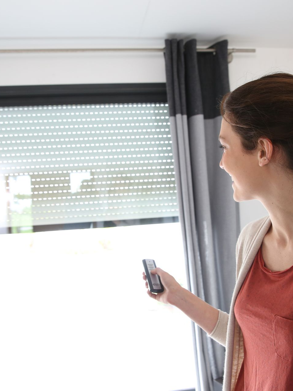 Une femme souriante actionne la télécommande d'un store enrouleur, à côté de rideaux et d'une fenêtre.