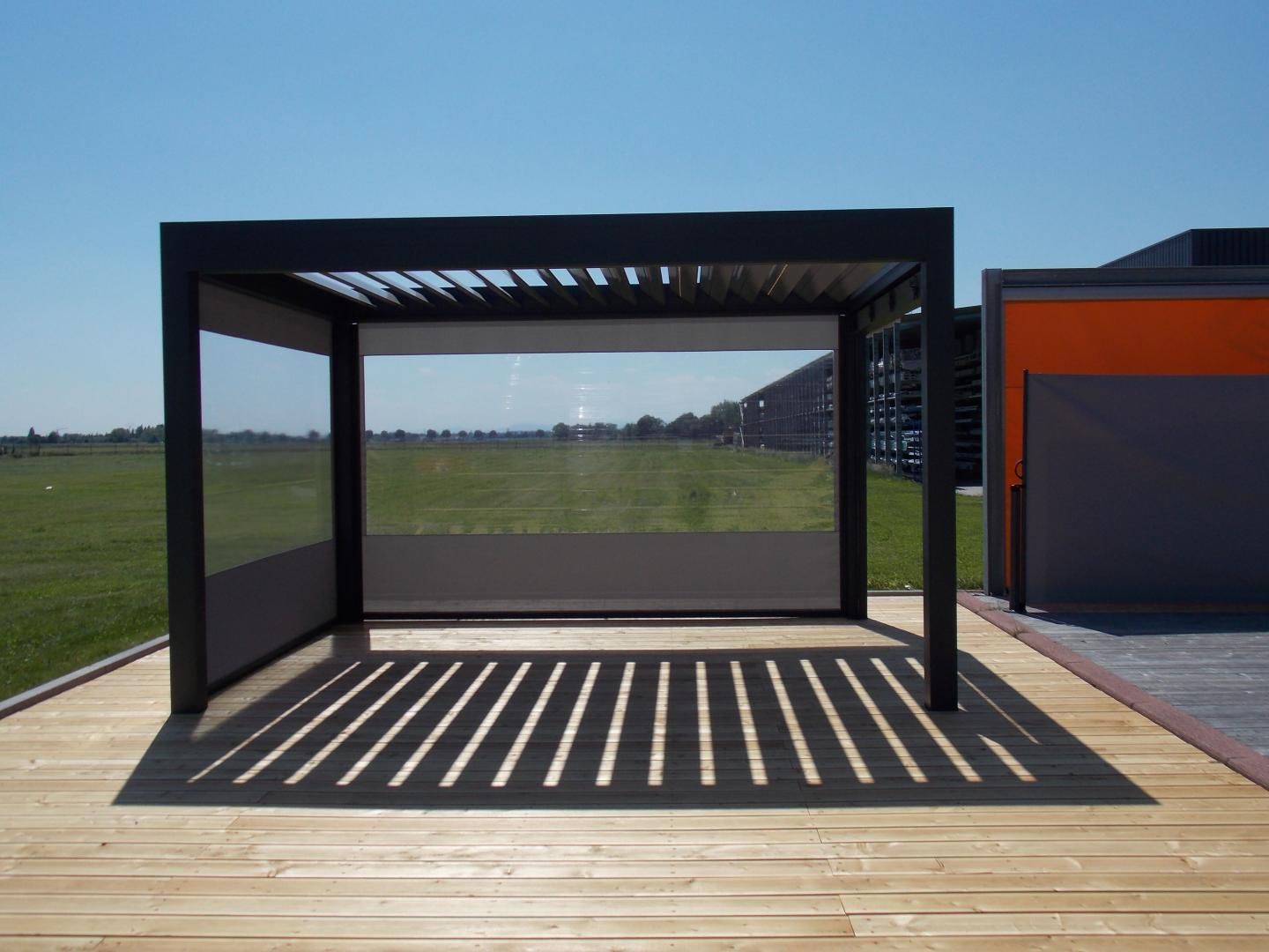 Pergola noire sur terrasse en bois projetant une ombre rayée, avec écrans latéraux partiels se détachant sur un ciel bleu.