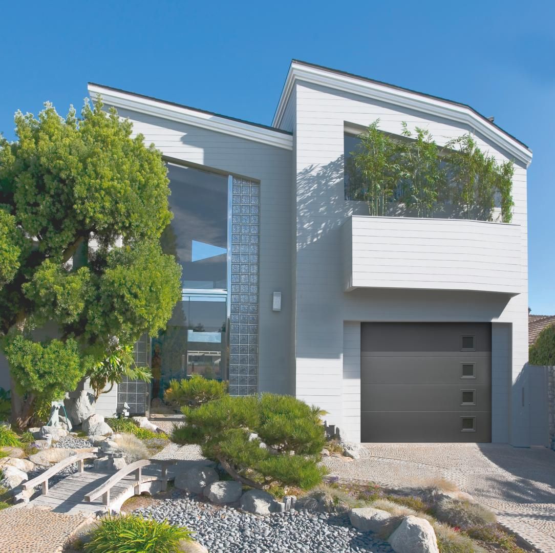 Maison moderne à deux étages avec porte de garage grise, grandes fenêtres et jardin paysager.