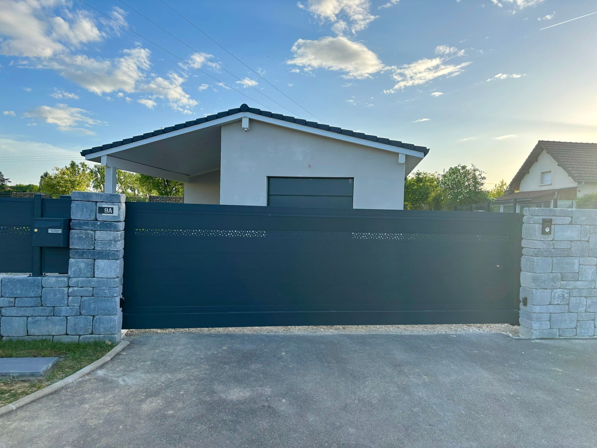 Portail coulissant gris foncé devant une maison claire avec un abri de voiture ; ciel bleu.