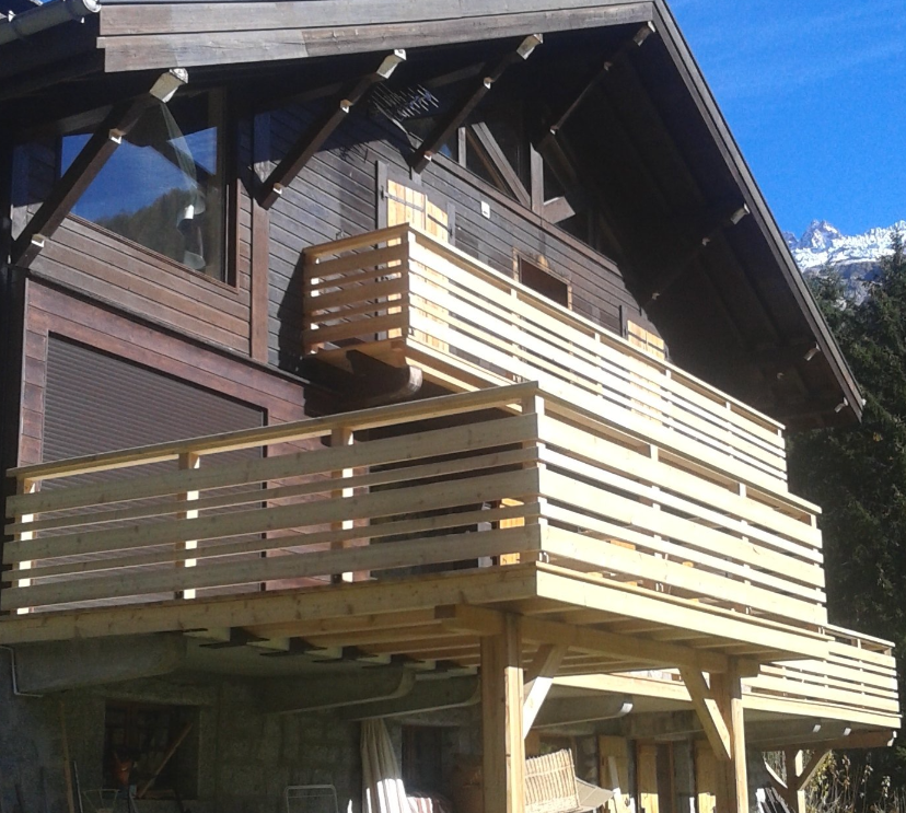 Maison en bois avec une grande terrasse enveloppante et une balustrade, située à flanc de montagne ensoleillé.