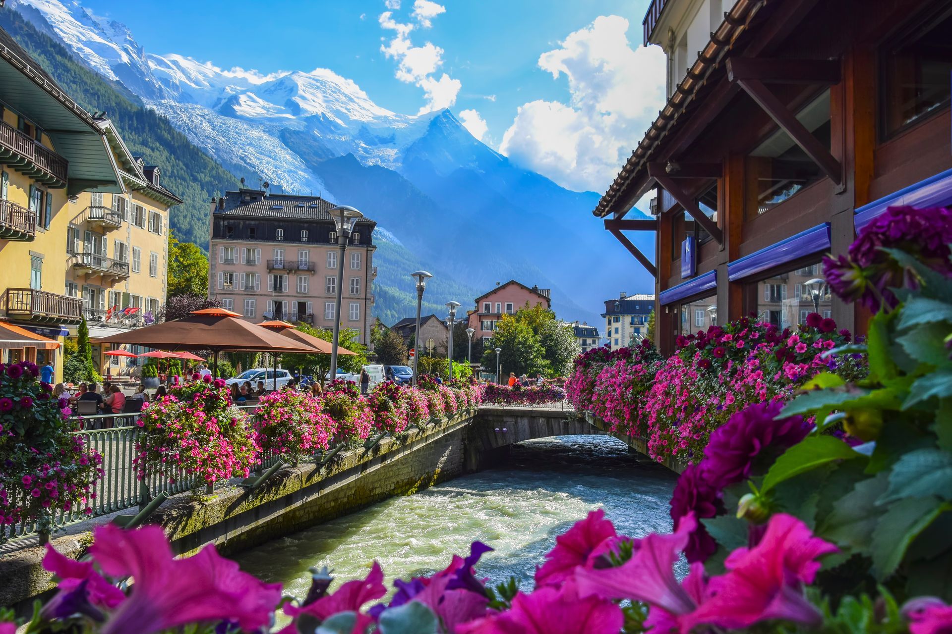 Village coloré au bord de la rivière, avec un canal bordé de fleurs et des montagnes enneigées sous un ciel d'un bleu éclatant