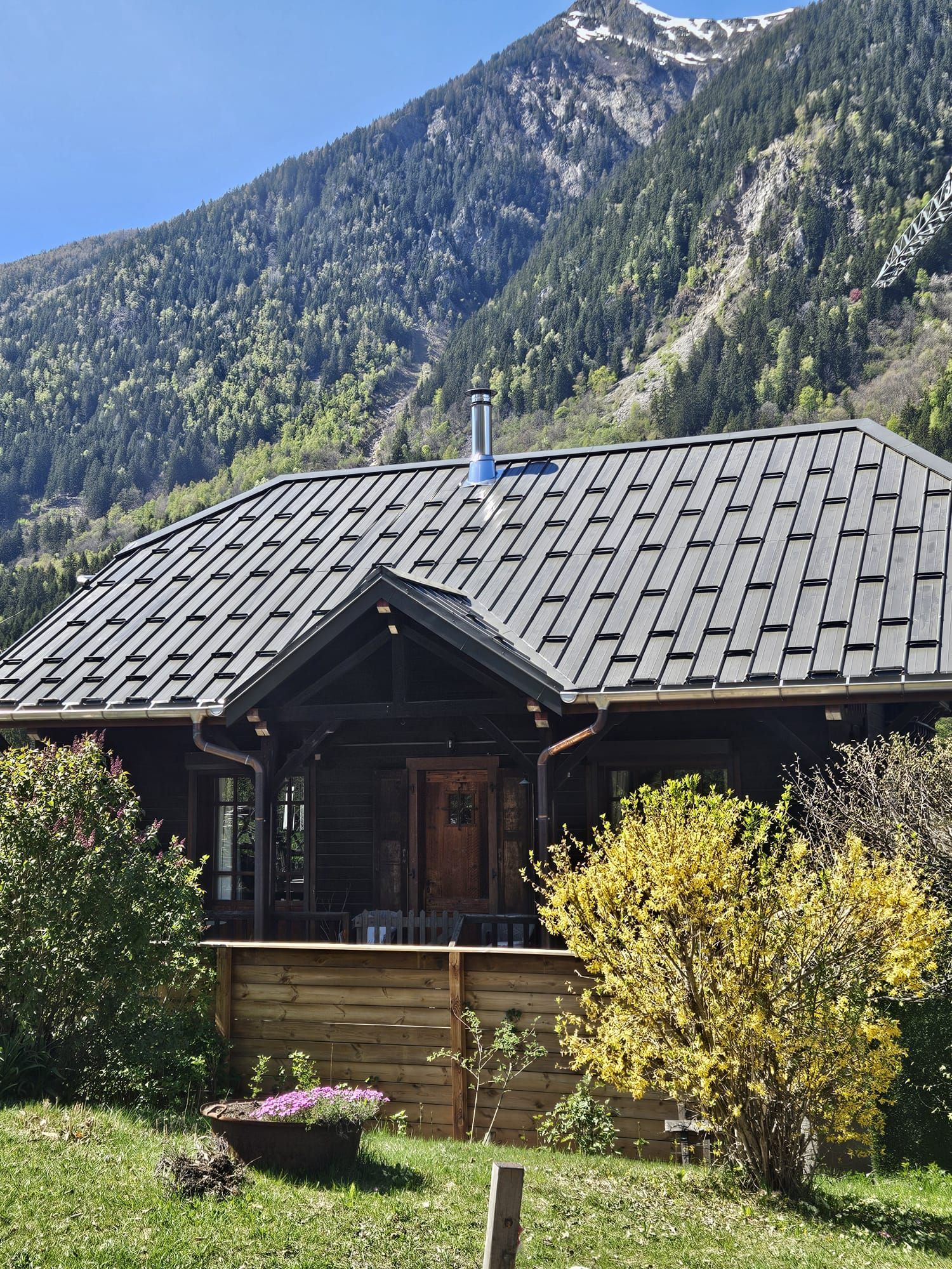 Cabane en bois au toit sombre devant des montagnes boisées sous un ciel bleu clair