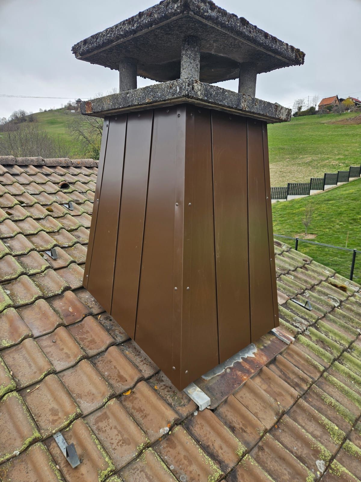 Cheminée brune sur un toit de tuiles, coiffée d'un petit chapeau, à côté d'une colline herbeuse.