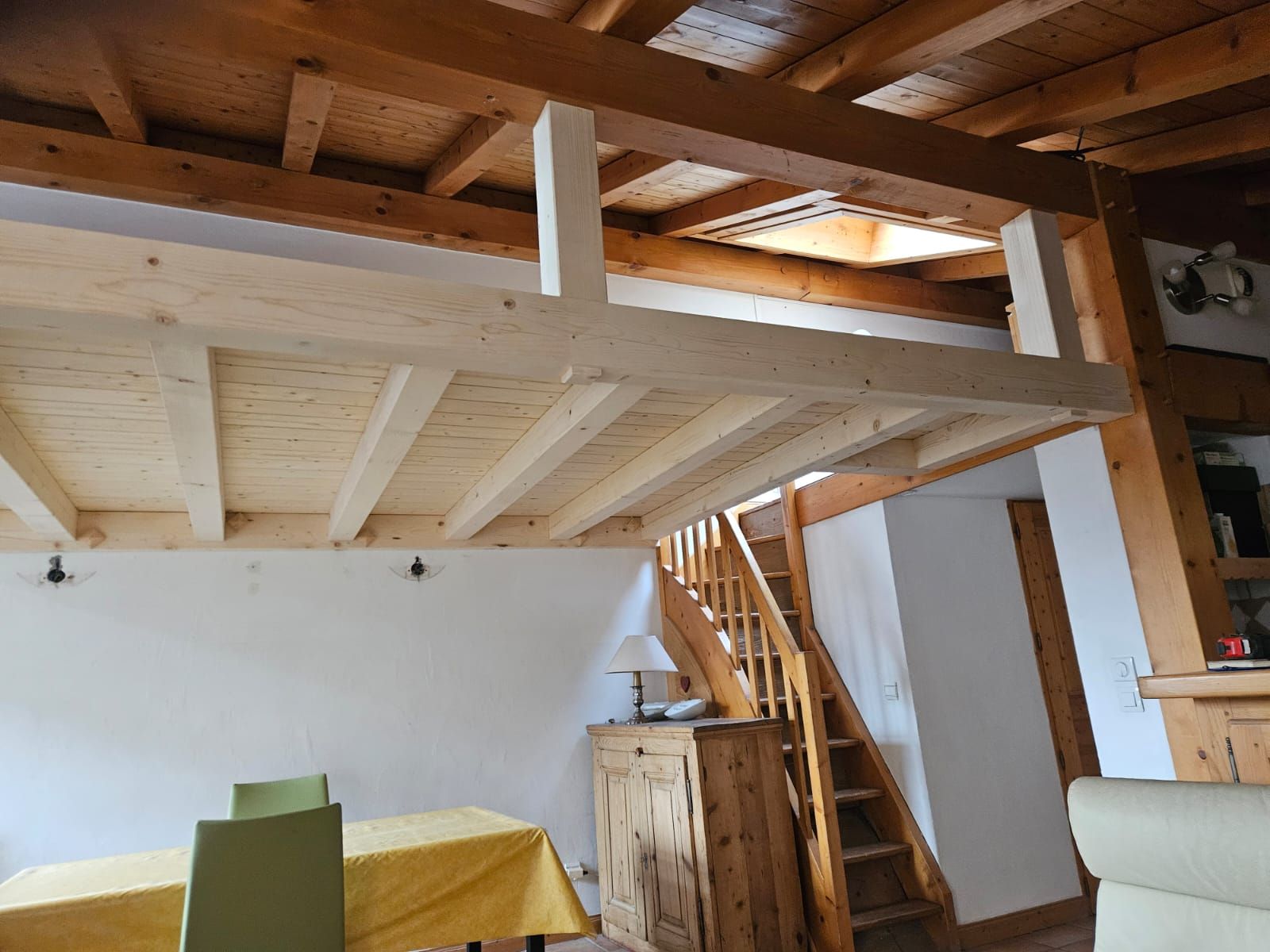 Pièce mansardée avec poutres apparentes, escalier et table à manger sous un plafond blanc incliné.