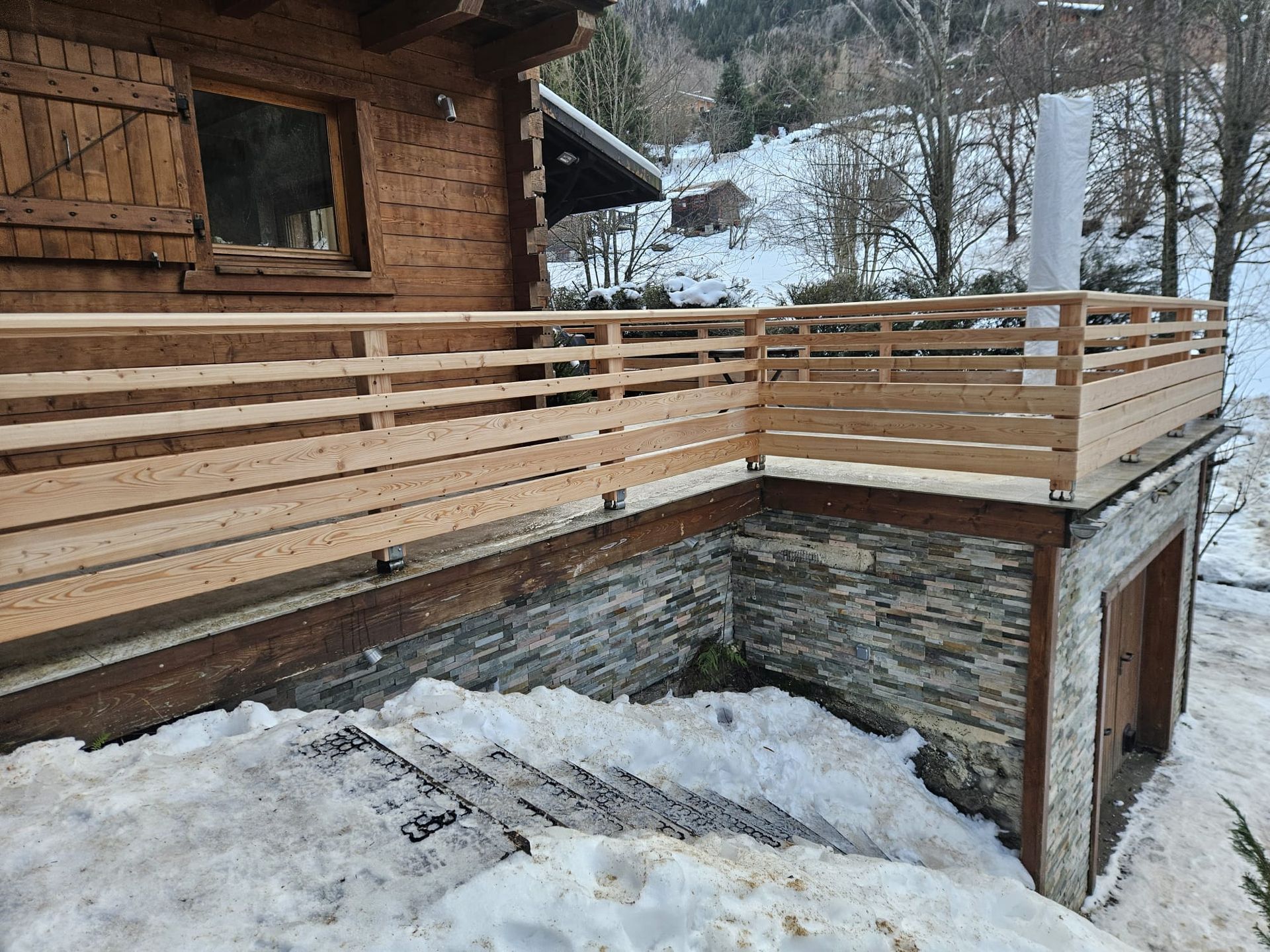 Terrasse en bois d'un chalet avec rambardes horizontales donnant sur une cour en pente enneigée en hiver.