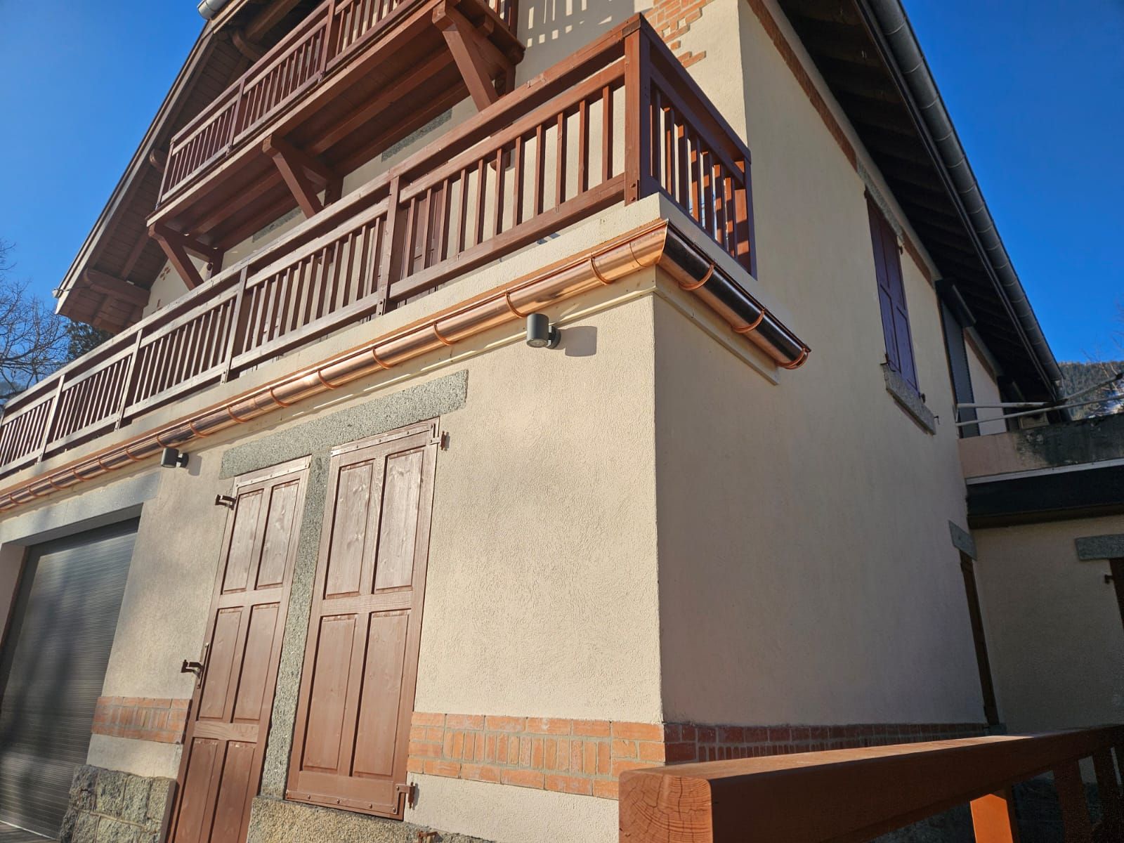 Ancien bâtiment beige avec balcon en bois et portes à volets, vu d'un angle bas sous un ciel bleu