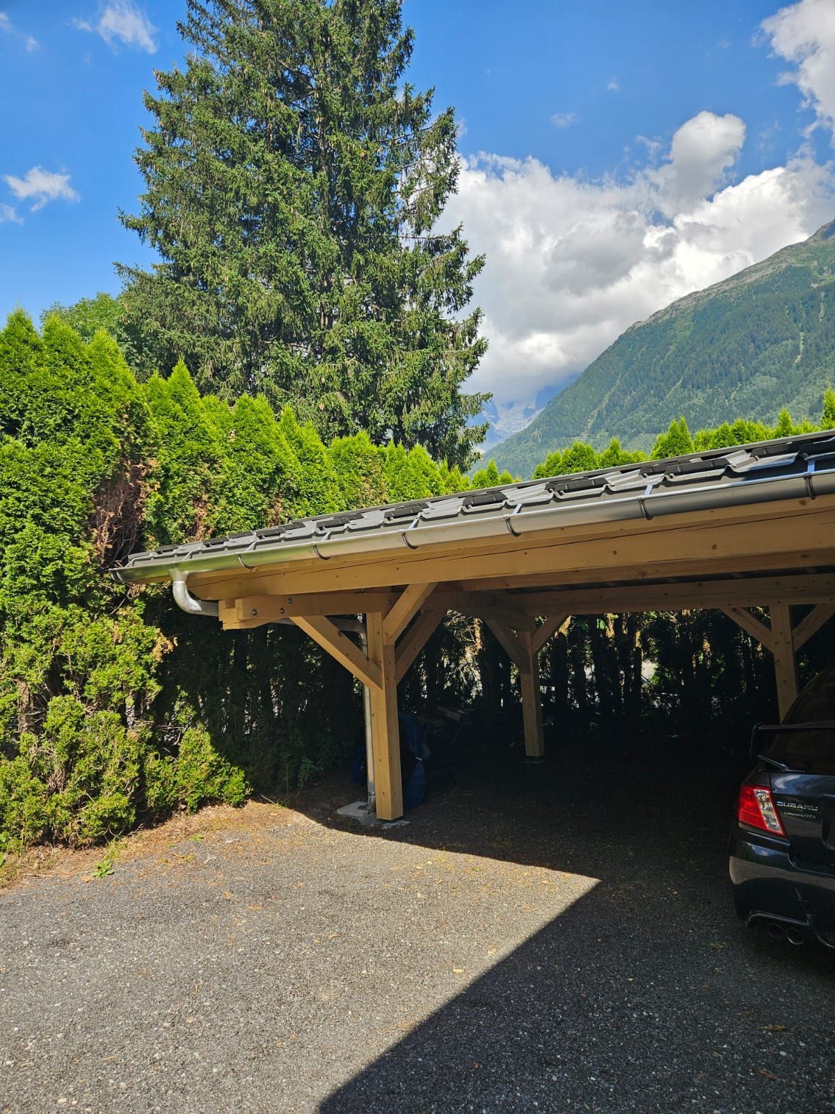 Allée sous un abri de voiture en bois, bordée de grands conifères et de montagnes par une journée ensoleillée