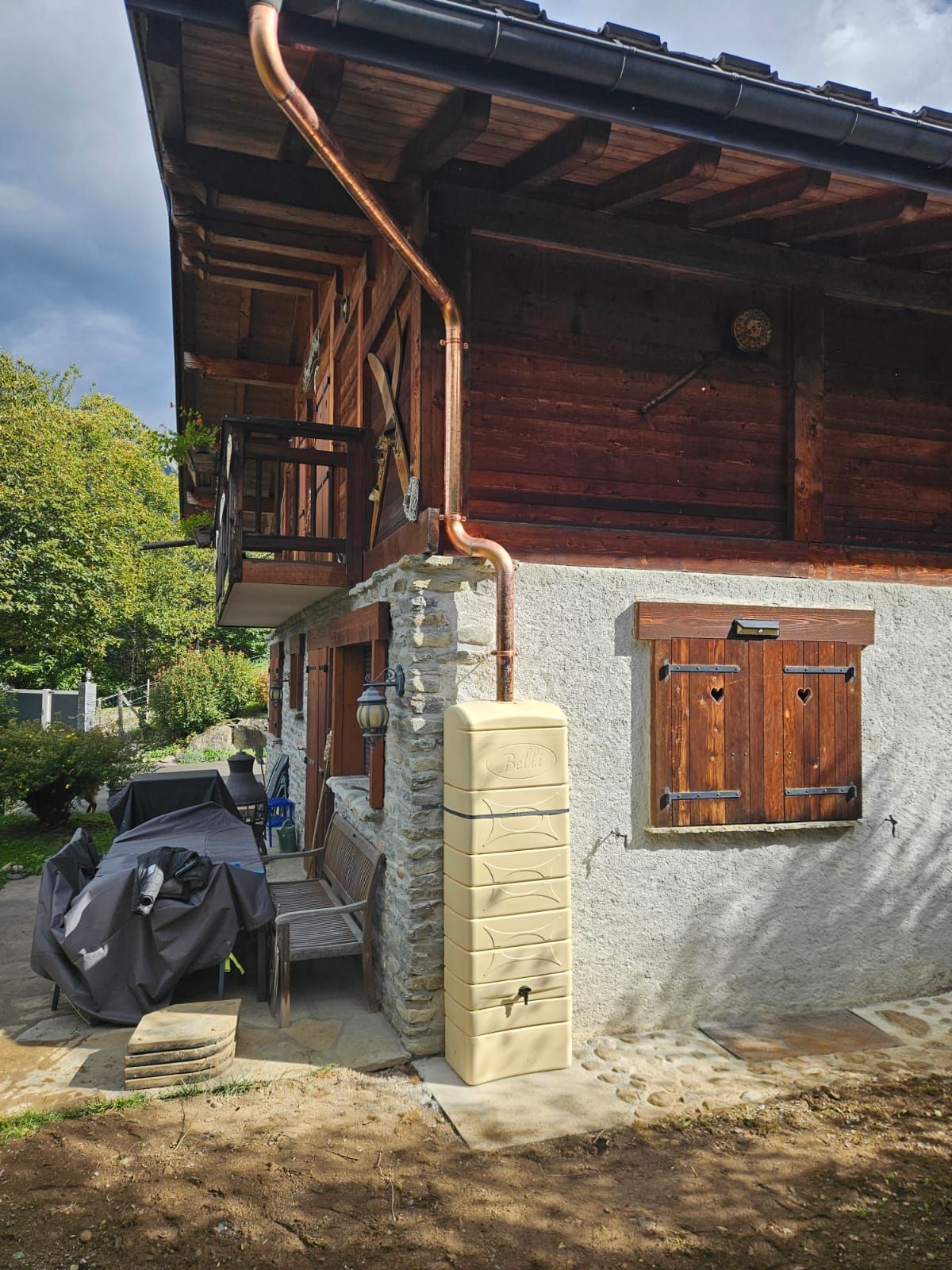 Extérieur rustique d'une maison en bois avec un muret en pierre, une fenêtre à volets et un scooter garé sous l'avant-toit.