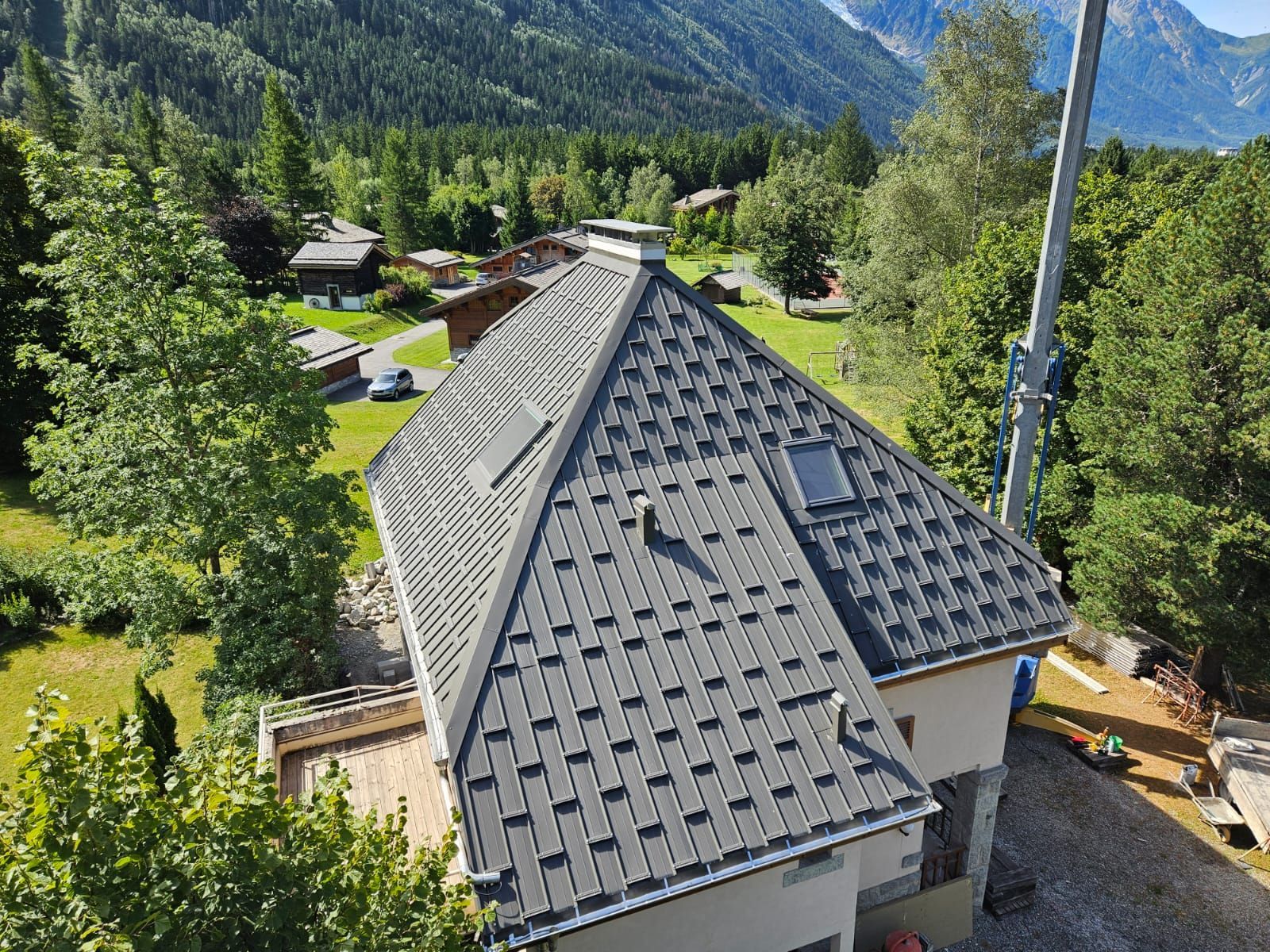 Une maison au toit gris dans une vallée montagneuse verdoyante, avec des arbres et un flanc de colline boisé au-delà.