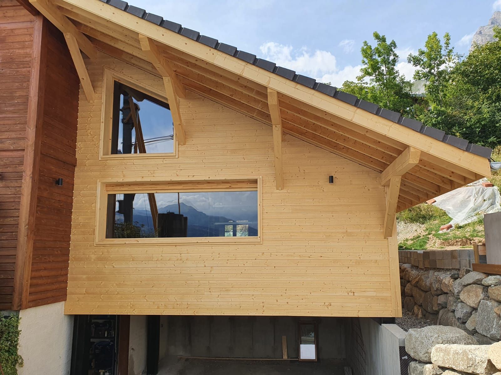 Façade de maison en bois avec toit en pente, grandes fenêtres et mur de soutènement en pierre