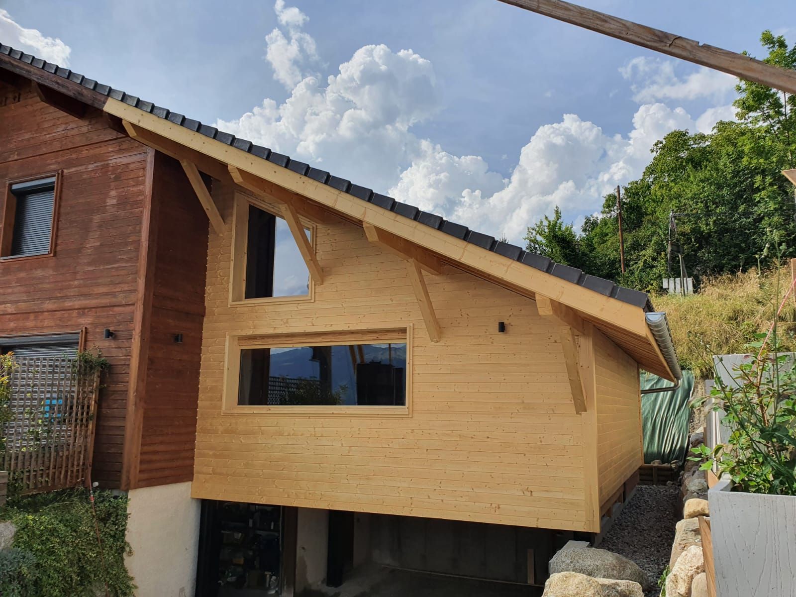 Nouvelle extension de maison en bois en construction à côté d'un bâtiment en briques, avec un toit en pente et un ciel nuageux