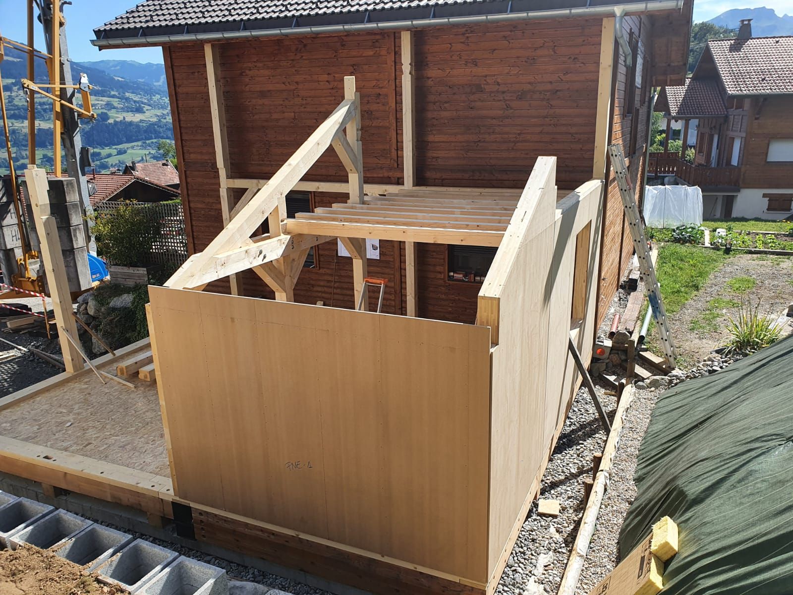 Petite structure en bois en construction au bord d'une route, avec des poutres de toit apparentes et des murs inachevés.