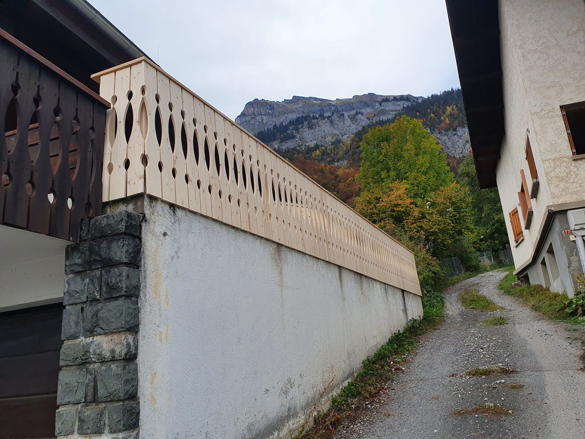Chemin étroit en montée entre des bâtiments, avec un mur de pierre et la montagne en arrière-plan sous un ciel nuageux.