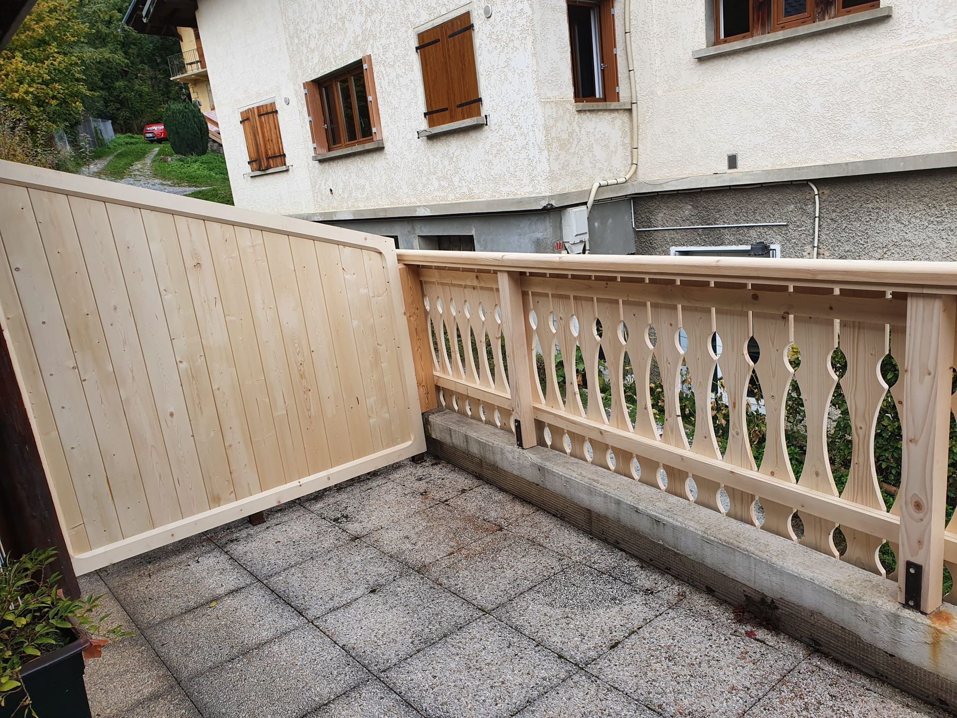 Petit patio carrelé avec un portail en bois crème et une balustrade décorative jouxtant un bâtiment blanc