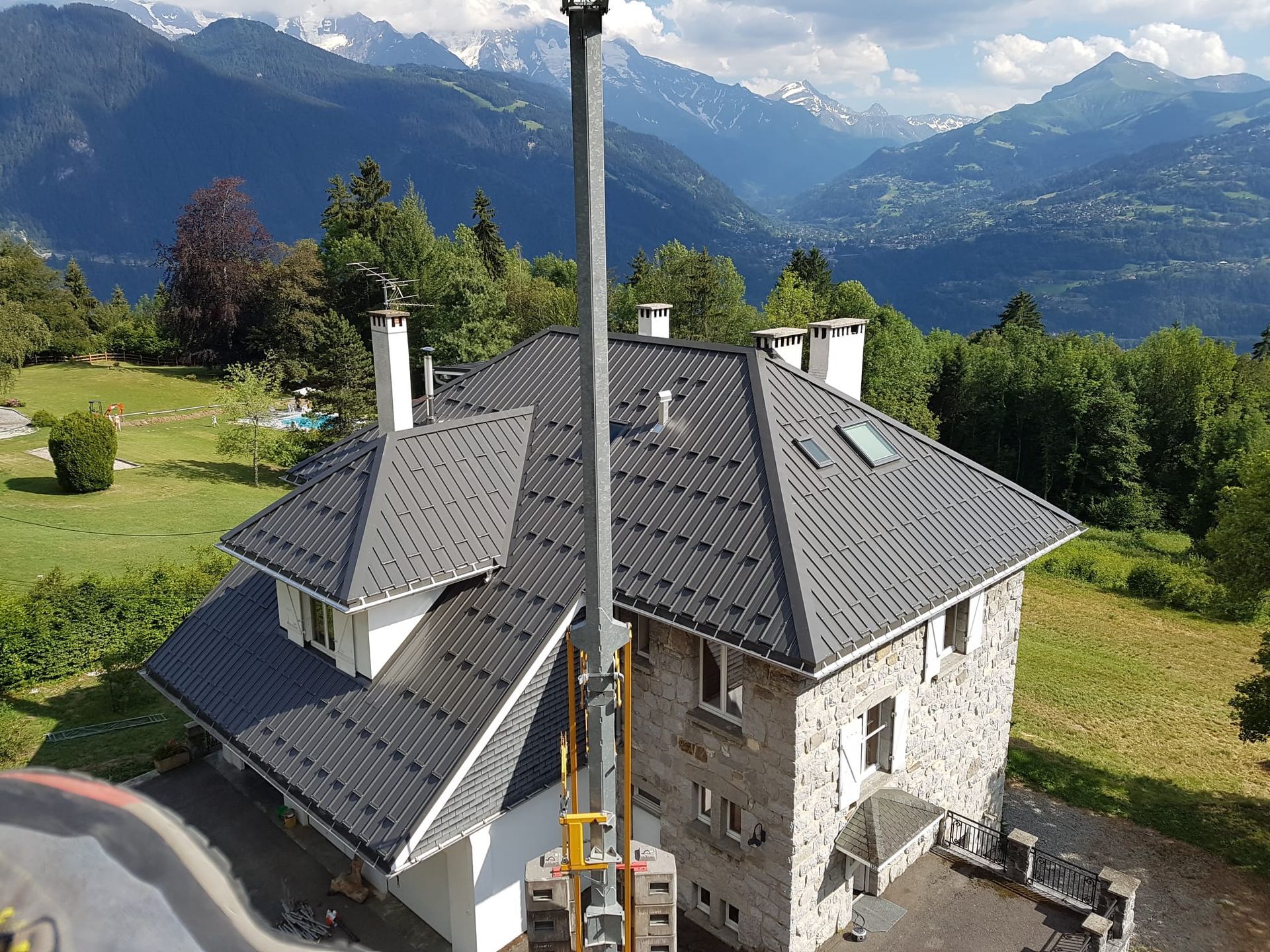 Maison en pierre au toit gris, à flanc de montagne, entourée de champs verdoyants et de montagnes en arrière-plan.