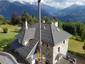 Maison en pierre au toit gris, à flanc de montagne, entourée de champs verdoyants et de montagnes en arrière-plan.