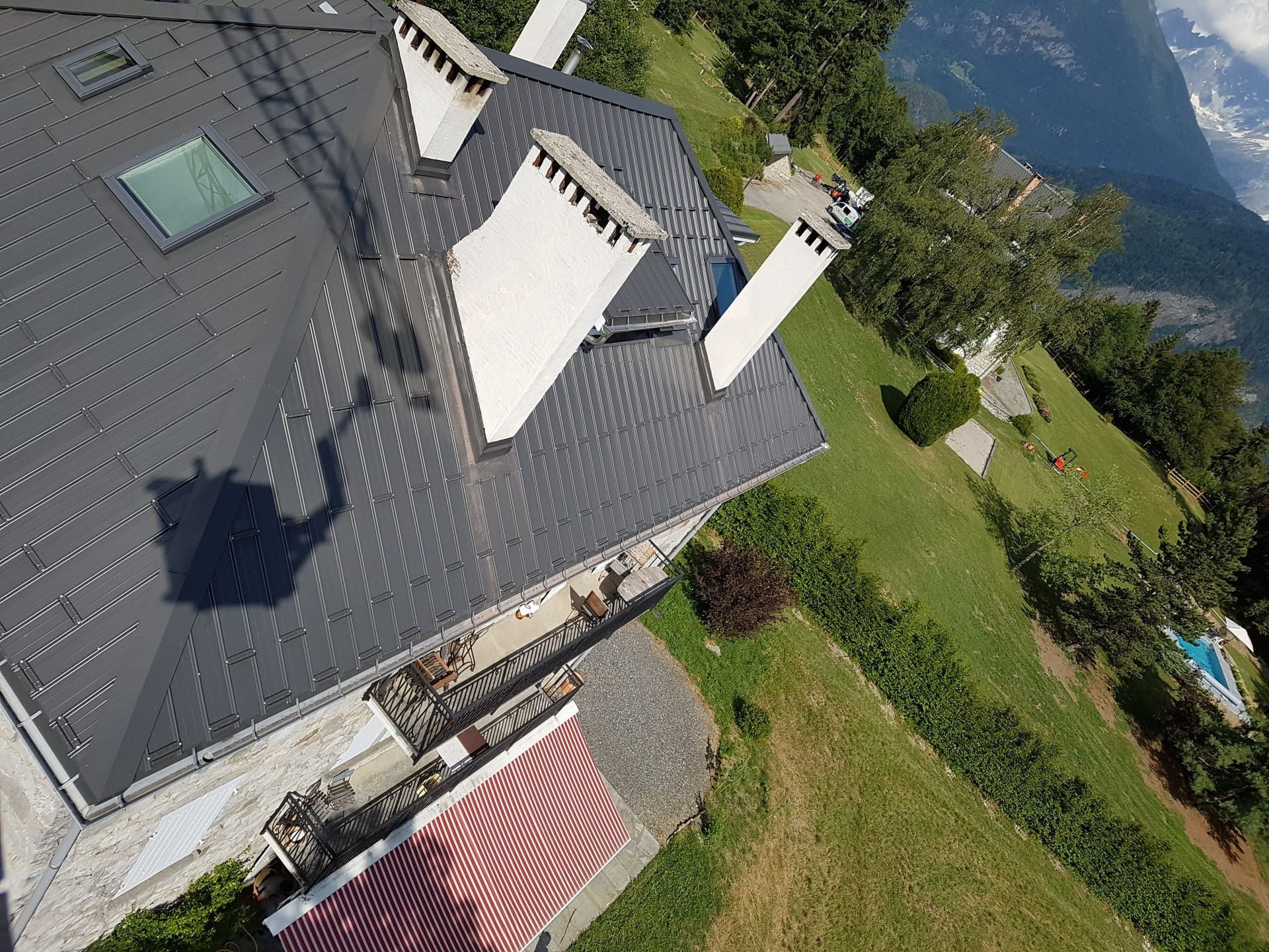 Vue aérienne d'une maison au toit gris située à côté d'une colline verdoyante et d'un lac.