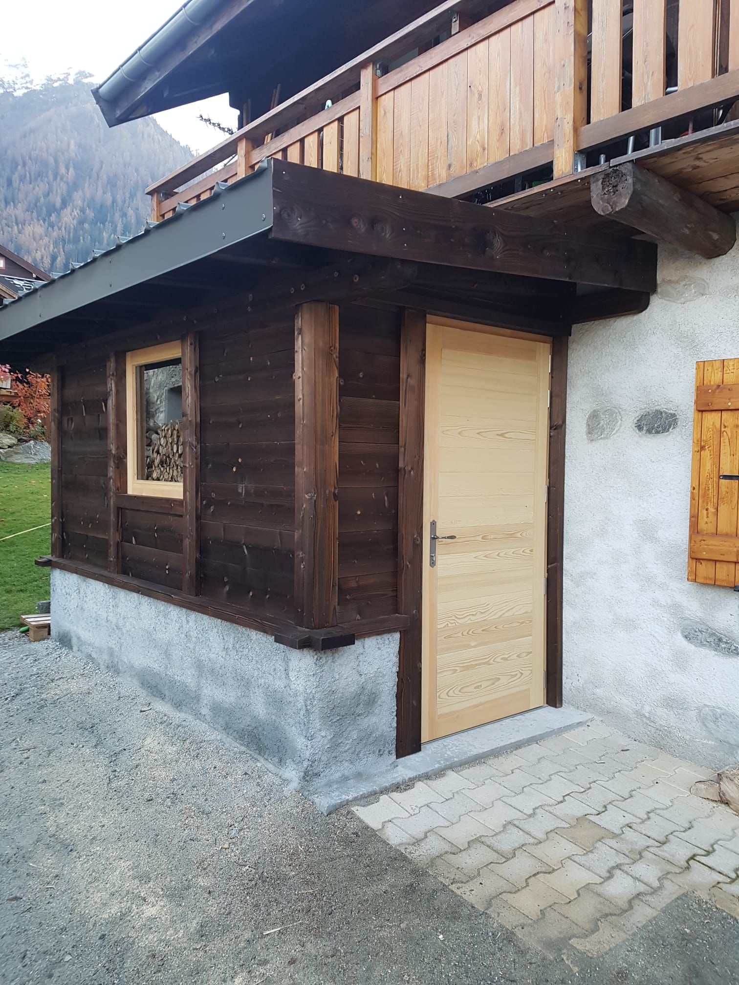 Petite entrée rustique en bois avec une porte beige, donnant sur un chemin de pierre au bord d'une route de gravier.