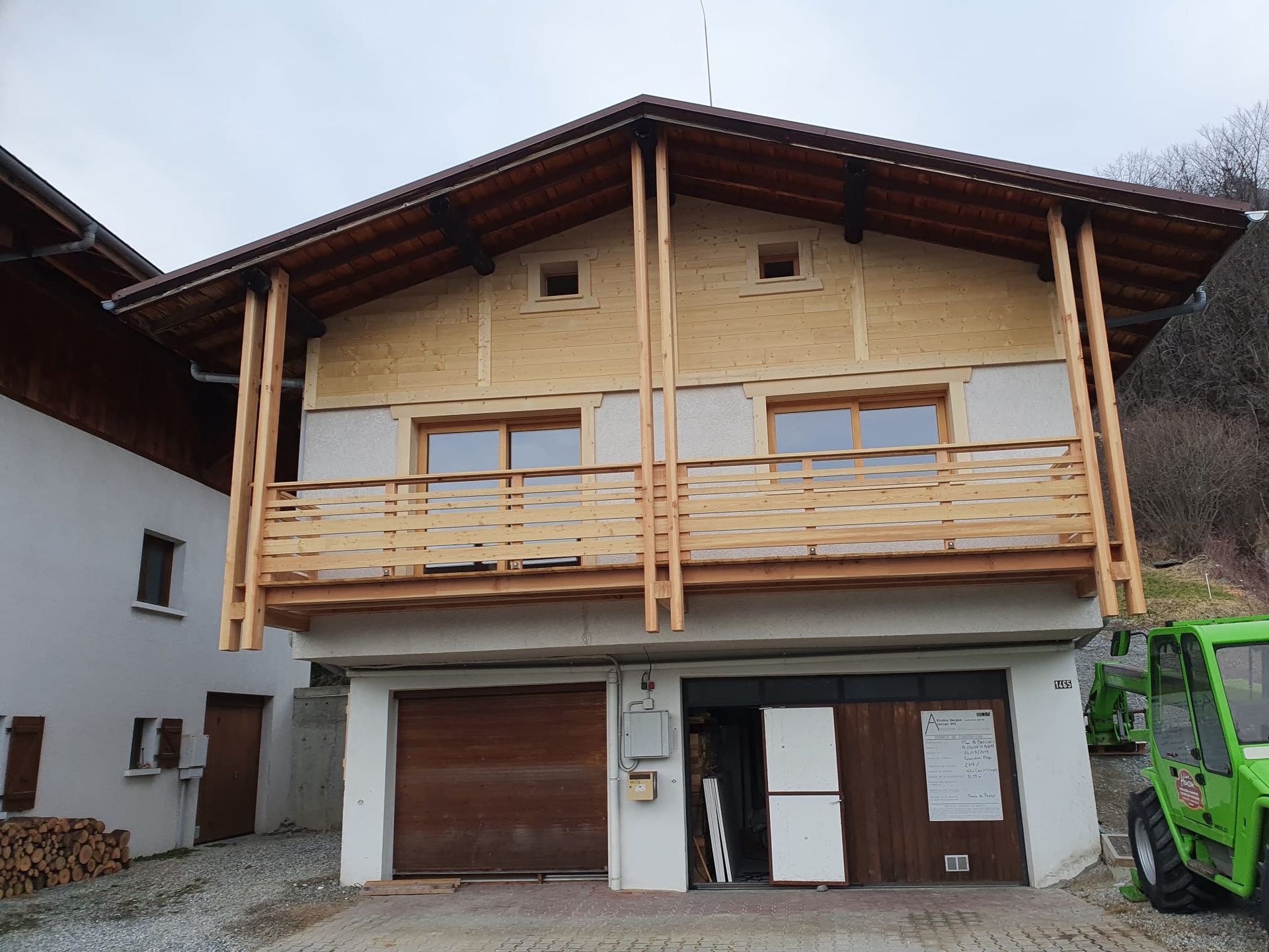 Maison de style chalet en bois avec balcon, garage et véhicule vert garé à côté.